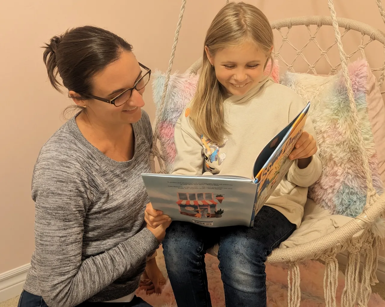 Izzy, wearing a beige sweater and jeans, sitting on a swing while holding a book beside her mother, Lindsey, wearing a grey top and jeans.