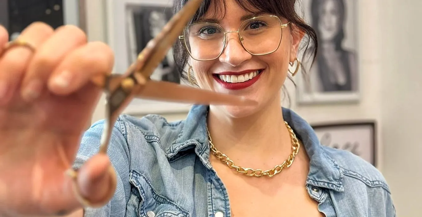 Jess, wearing a black dress and blue jacket, holding a pair of scissors.