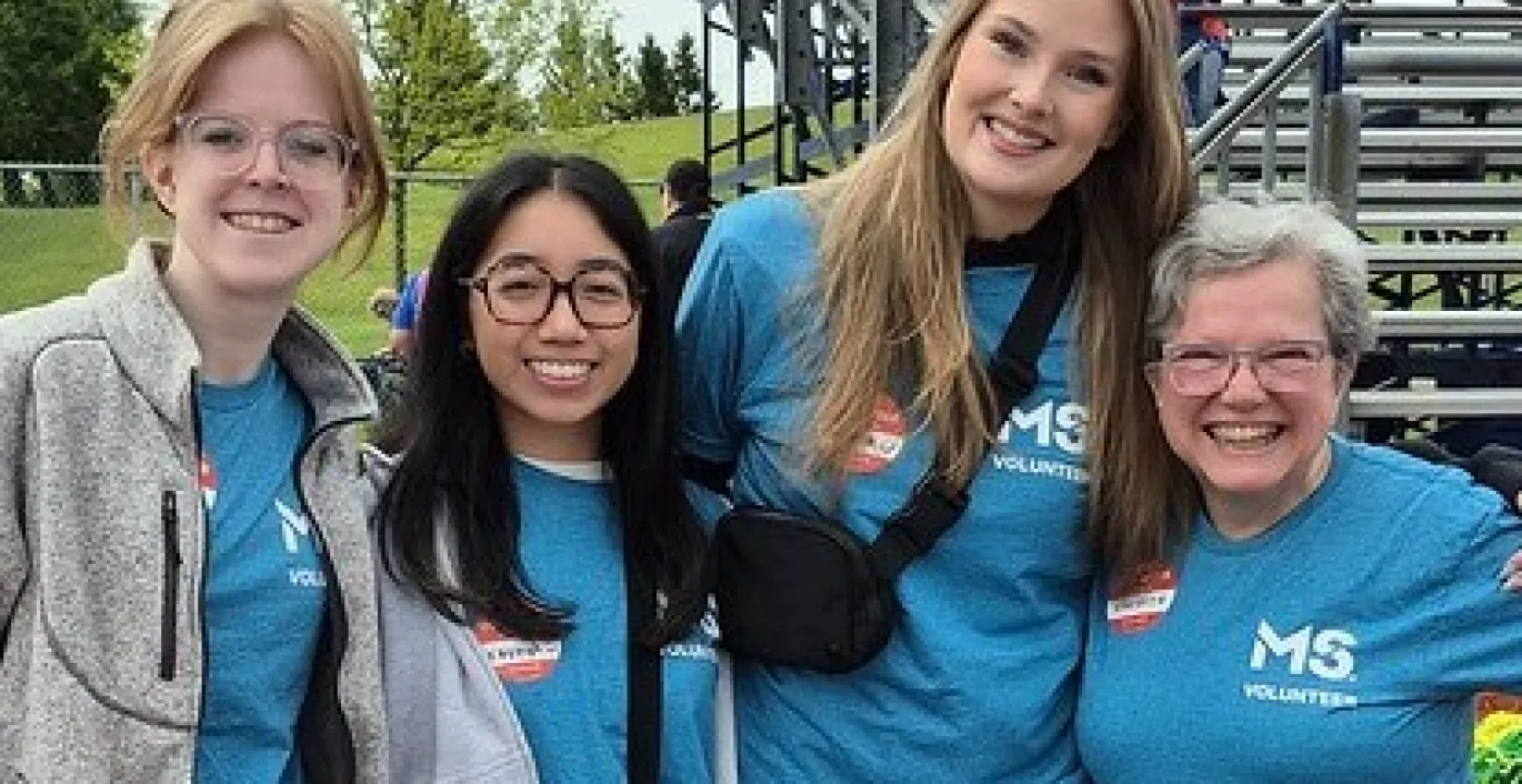 Four smiling women outdoors, three wearing blue MS charity shirts, at an outdoor event.