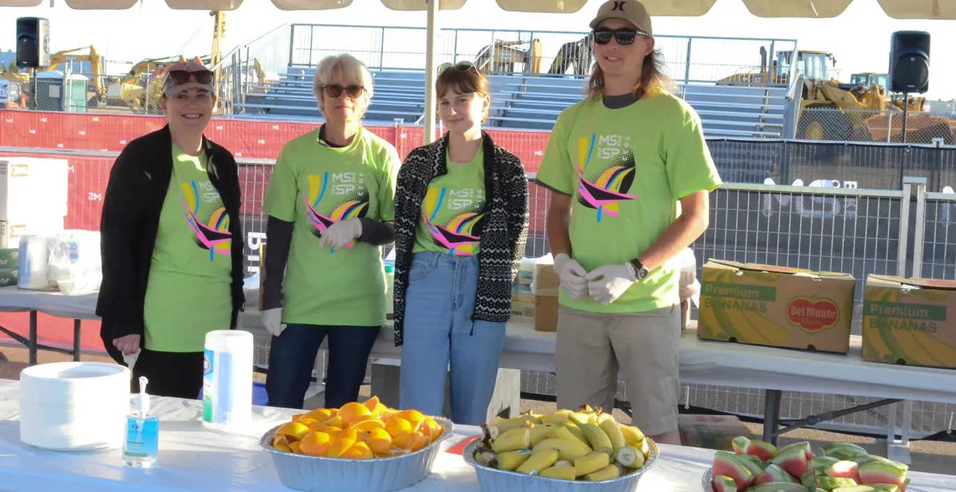Four volunteers in green T-shirts standing behind a table with fruit at an outdoor event.