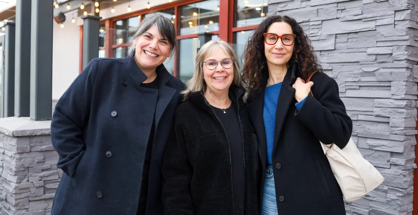 Three women smiling together outdoors, wearing dark coats on a cloudy day.