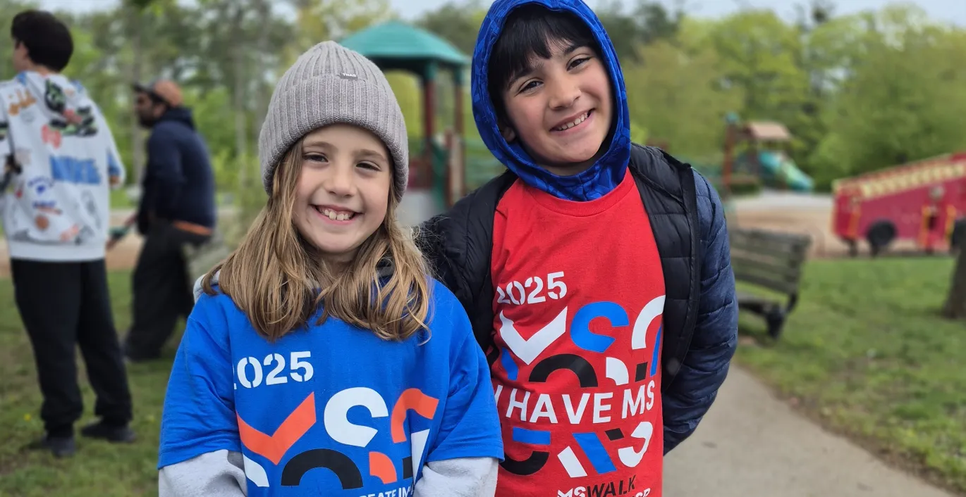 Two smiling children in numbered race bibs at an outdoor charity walk event.