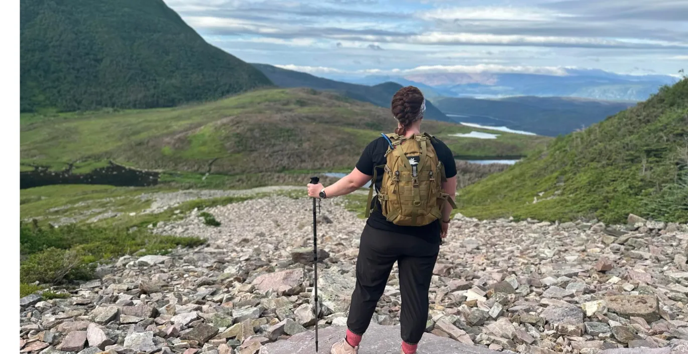 Janice with backpack and trekking pole standing on rocky mountain trail overlooking a valley.
