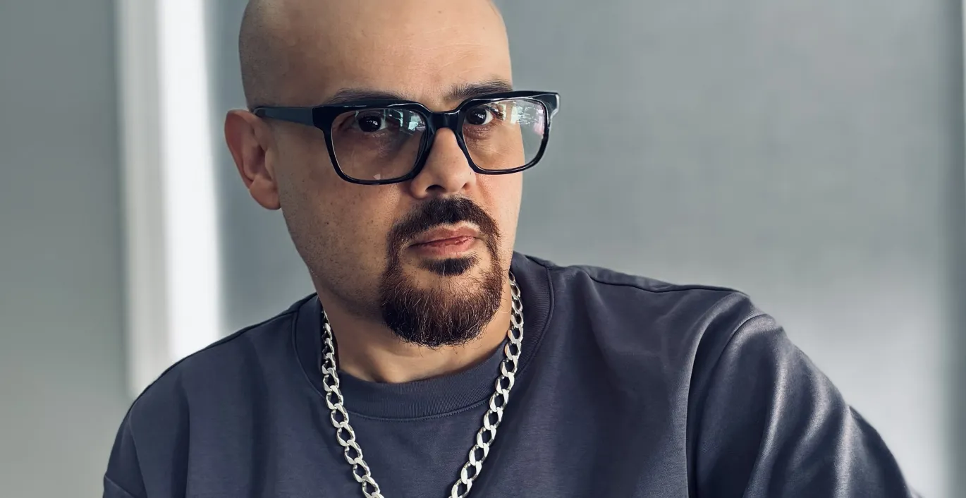 Miguel in gray t-shirt and chain necklace, wearing glasses, seated indoors.