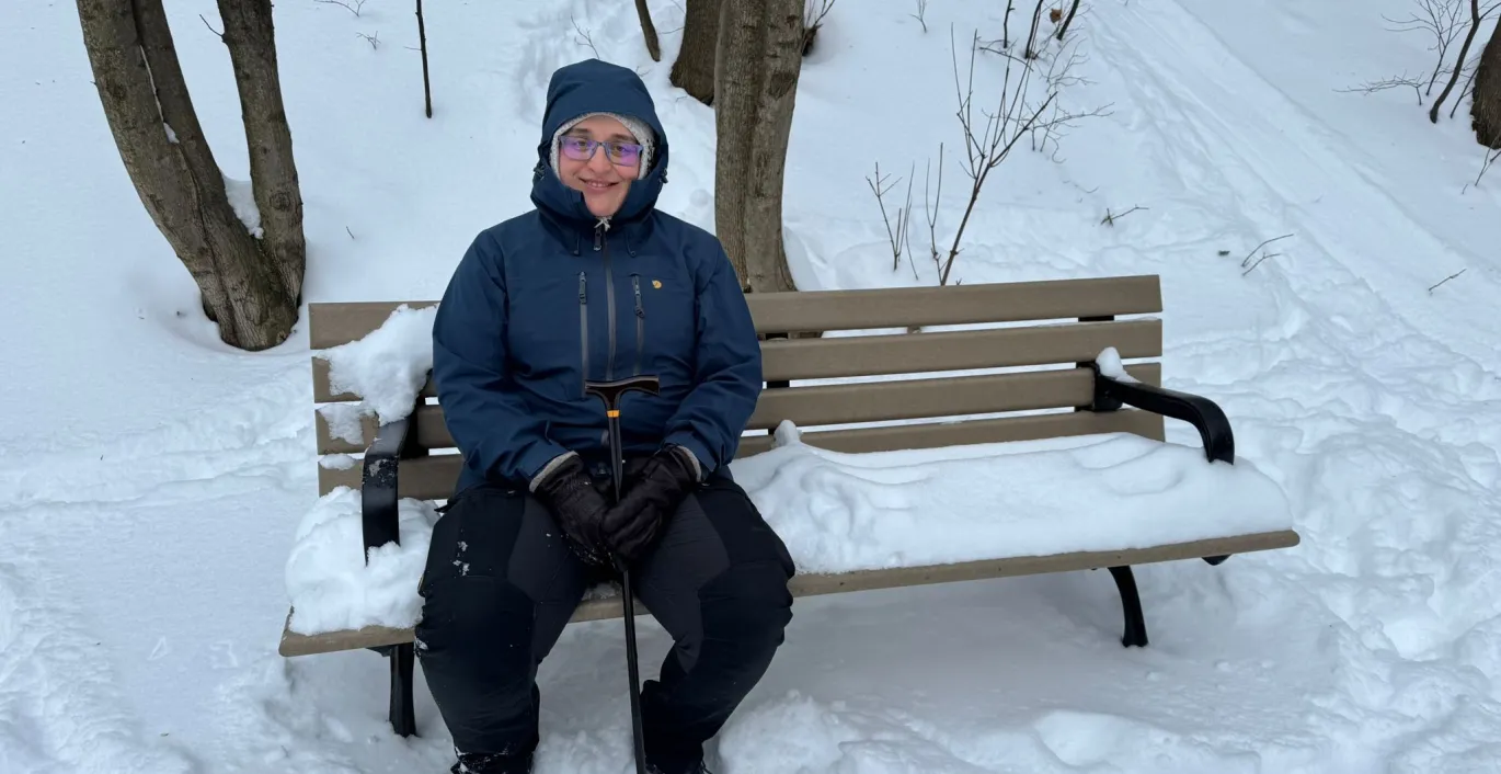 Ksenia in winter gear sitting on a snow-covered park bench outside.