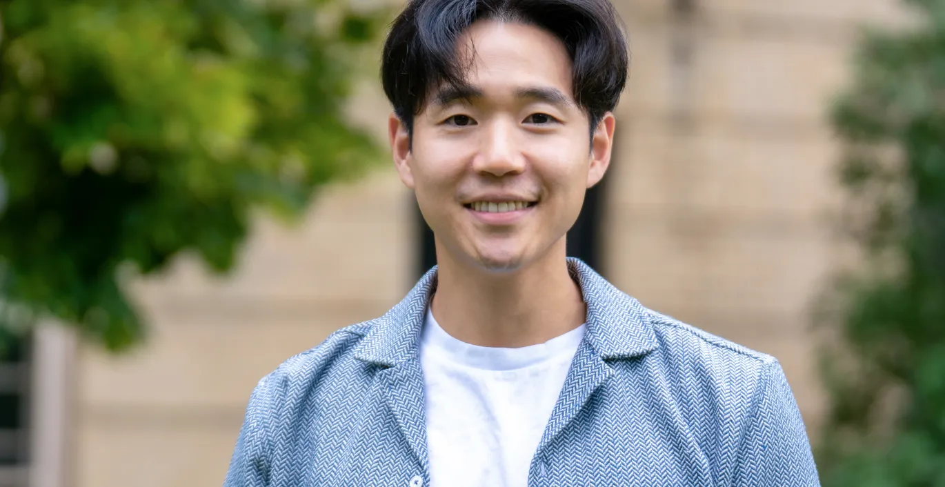 Dr. Paul Yoo is wearing a white t-shirt with a light blue blazer. He's smiling and standing in front of a building and trees.