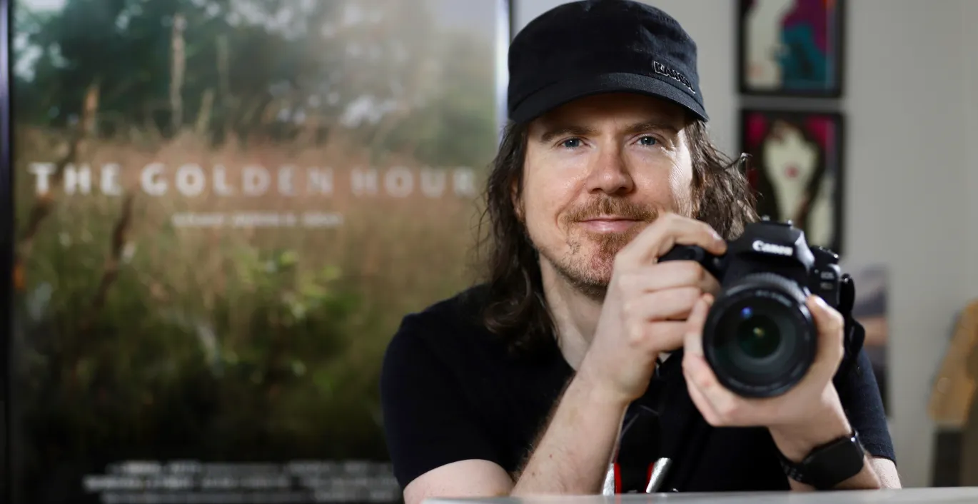 Jason wearing a black t-shirt and hat, holding a camera with a poster that reads "The Golden Hour" behind him.