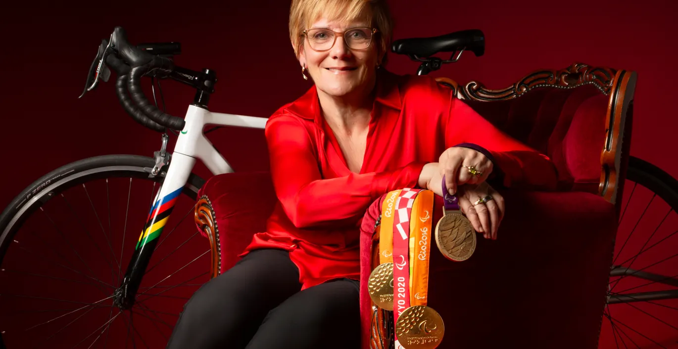 Carol in a red top and black pants sitting in a red armchair, holding medals, with a road bike behind her. 