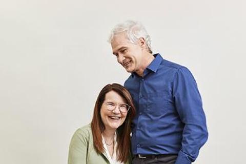 A woman in wheelchair with long red hair and glasses wearing a sage green blazer embraces a man with a blue shirt and white hair.