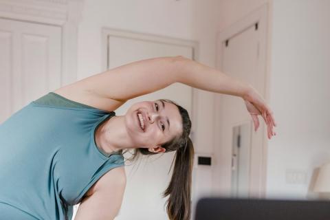 A woman with a long, brown ponytail wearing workout clothing does a side bend while in front of her latop.