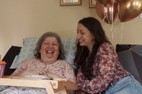Stephanie, in a floral blouse, smiling at her mother, Carm, who is lying in bed in a pink pajama top with pink balloons in the background.