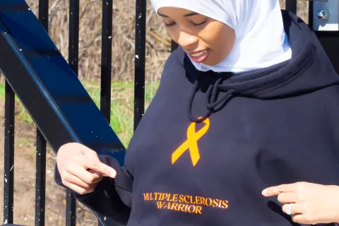 Najma sitting and looking down while pointing at black hoodie that reads 'Multiple Sclerosis Warrior.'