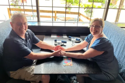 James, wearing a black shirt, and Yolanda, wearing a blue dress, sitting at a restaurant booth and holding hands.