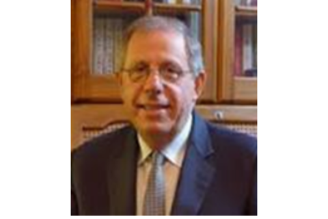 Portrait of Dr. Anthony Feinstein, a middle-aged man with short gray hair and glasses, wearing a dark suit and a blue tie. He is smiling and sitting in front of a wooden bookshelf filled with books.