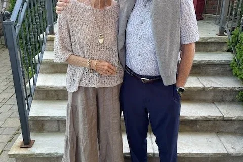 Jan and Tom, smiling in front of some steps outside. Jan is wearing a tan coloured shirt and skirt and Tom is wearing a light blue shirt, grey sweater around his shoulders, and blue pants..