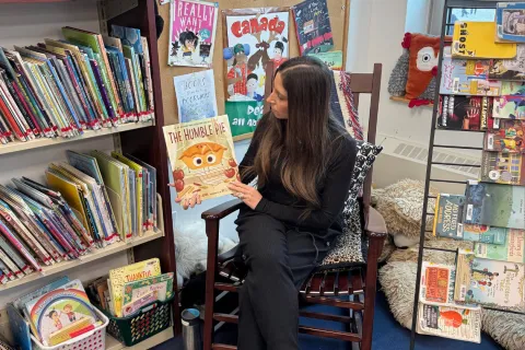 Jaclyn, wearing a black top and black pants, sitting in a chair, holding up a book.