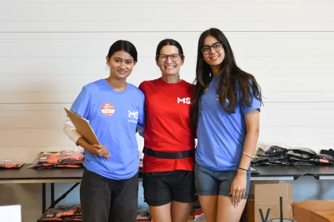 Three MS Canada volunteers stand smiling together indoors during event registration. One volunteer holds a clipboard, while tables behind them are filled with folded volunteer T-shirts and event supplies.