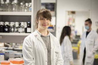 MS Researcher Dr. Catherine Larochelle, a woman with brown hair wearing a lab coat and gray shirt in a lab setting.