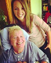 A woman with long brown hair smiles and stands behind her mother, who has short white hair and is wearing a blue button up shirt. She's smiling too.