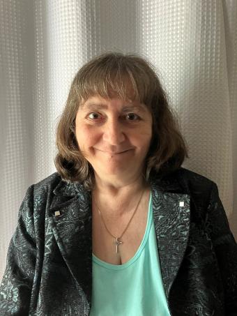 A woman with short brown hair and bangs is wearing a teal shirt with a dark blazer.