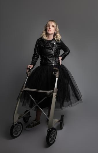 A woman with blond hair is wearing a leather jacket and tulle skirt. She is holding onto a walker and has her other hand on her hip.