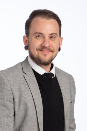A man with short brown hair is wearing a grey suit and black vest with tie.