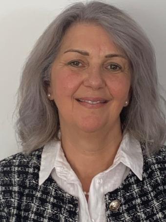 A woman with medium length grey hair is wearing a black and white tweed jacket and collared shirt.