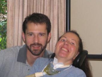 A man with brown hair and a short beard is next to a woman with long hair who has an oxygen mask around her neck.