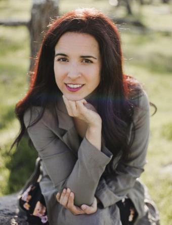 A woman with long dark hair is wearing a floral dress with a dark grey blazer.
