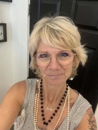 A woman with short blond hair is wearing round glasses and a grey tank top.