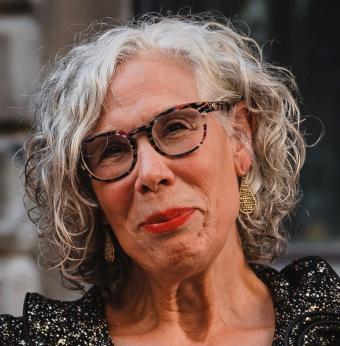 A woman with short, grey curly hair is wearing dark rimmed round glasses and red lipstick. She is smiling.