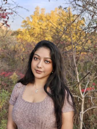 A woman with long brown hair is wearing a light brown t-shirt. She is in front of a backdrop of fall trees.
