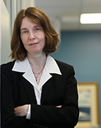 A woman with medium length red hair is wearing a black suit and white collared shirt. She leans on the wall with arms crossed.
