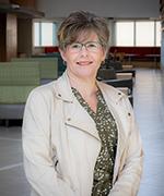 A woman with short brown hair is wearing glasses and a beige jacket with a printed shirt. She stands in front of a hallway filled with seating.