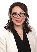 A woman with long, curly, dark hair is wearing black rimmed glasses and a beige suit. She smiles. 