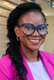 A woman with long dark hair is wearing round glasses with dark blue rims and a bright pink blazer. She is smiling.