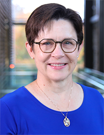 A woman with short brown hair is wearing round dark rimmed glasses and a royal blue shirt. She is smiling.