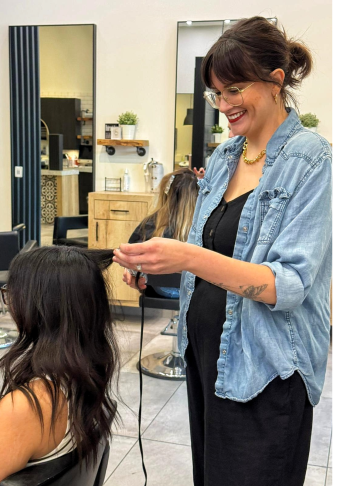 Jess, wearing a black dress and blue jacket, styling a client's hair.