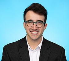 Man with short dark hair swept to the side smiling, wearing glasses and a dark blazer, against a light blue background.