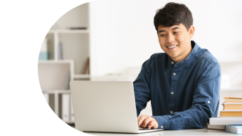 Young man in a blue shirt smiling while working on a laptop at a desk.