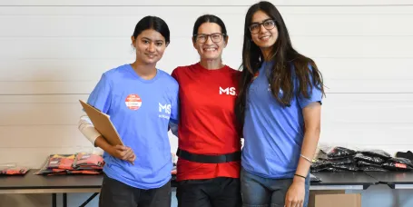 Three MS Canada volunteers stand smiling together indoors during event registration. One volunteer holds a clipboard, while tables behind them are filled with folded volunteer T-shirts and event supplies.