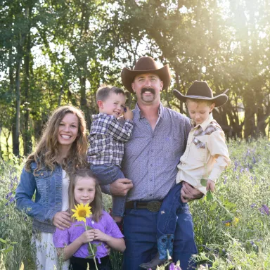 Family of five posing in a sunny wildflower meadow with lush green trees in the background.