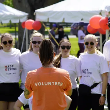 A volunteer in an orange shirt addresses a group wearing white "Make Strides" t-shirts at an outdoor event.