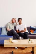 Two smiling men sitting together on a blue sofa in a bright living room.