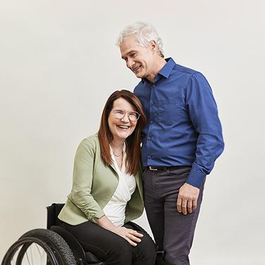 A woman in wheelchair with long red hair and glasses wearing a sage green blazer embraces a man with a blue shirt and white hair.