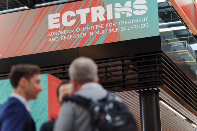 [A poster of ECTRIMS in focus, with people networking in the background]