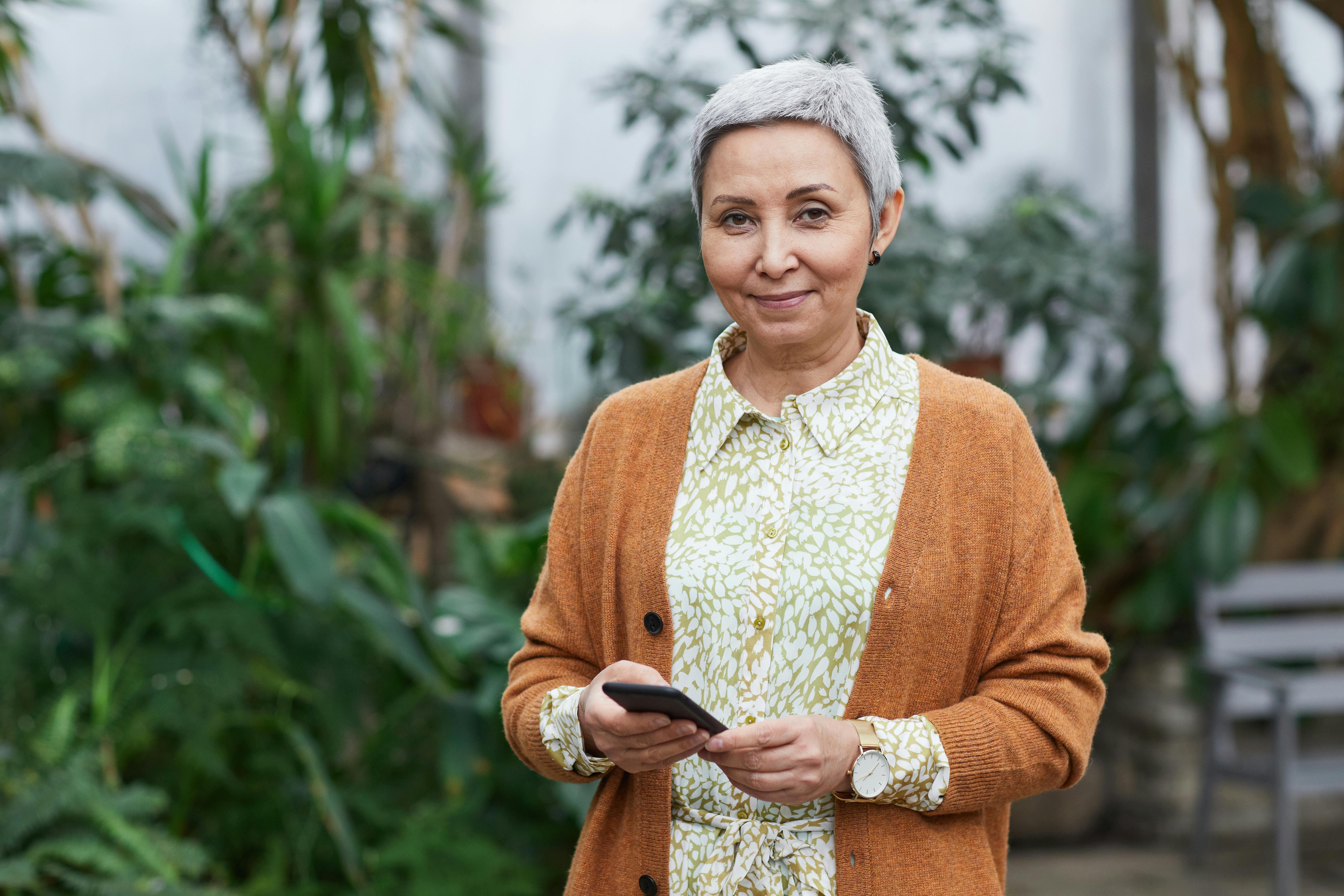 A woman with short grey hair is wearing a mustard coloured cardigan with a white and green floral shirt is holiding her cell phone. 