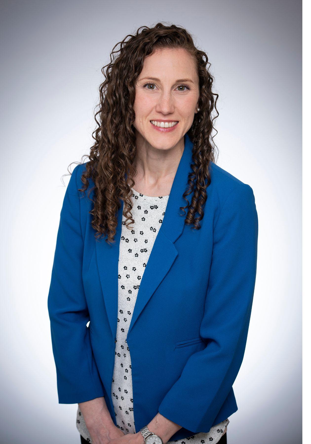 Laura is standing and smiling while wearing a royal blue blazer with a white and blue polka dot top underneath. She has curly brown hair and green eyes. 