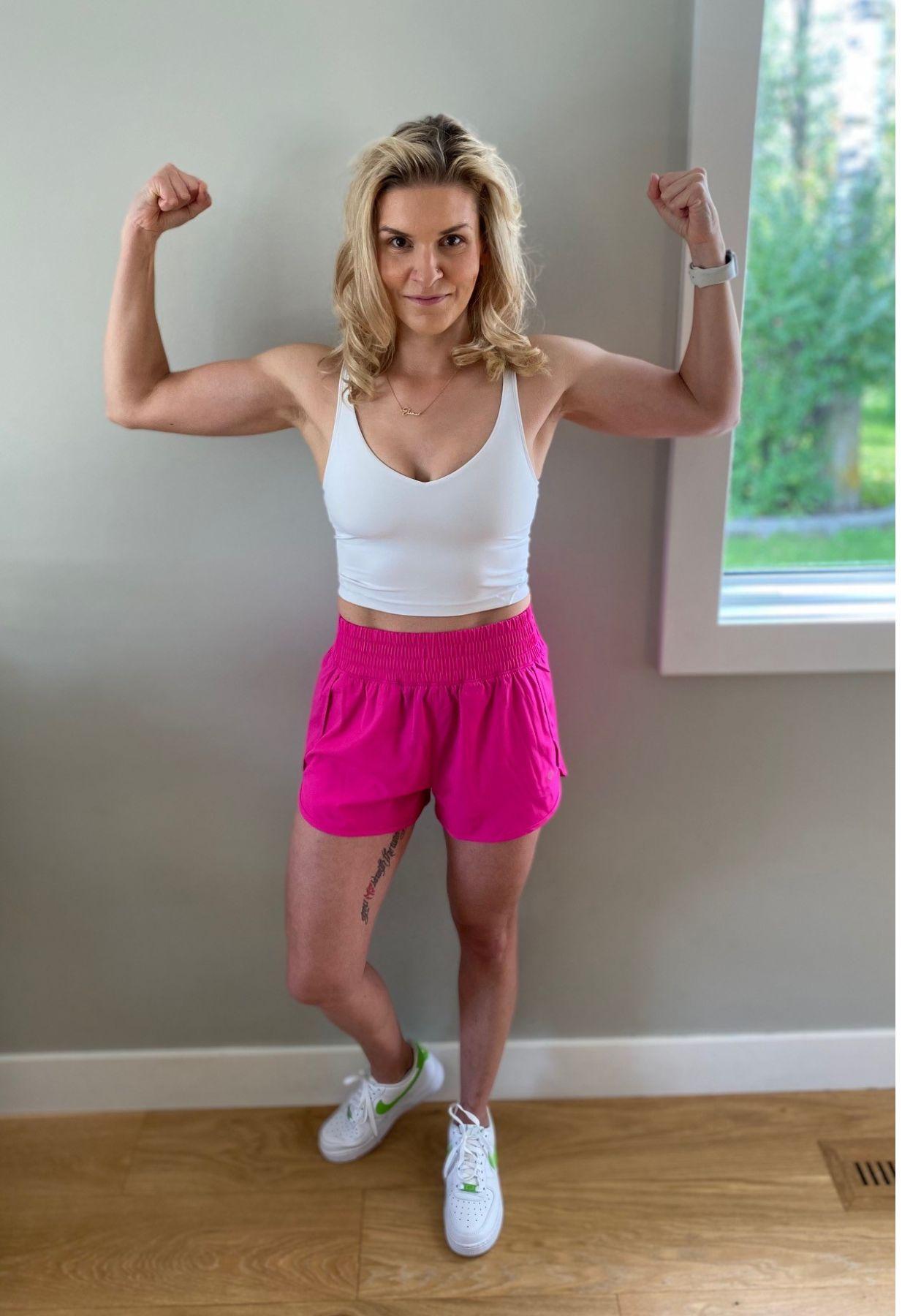 A woman, Patrycia, flexing her arm muscles, while wearing a white tank top with bright pink athletic shorts, and white sneakers.
