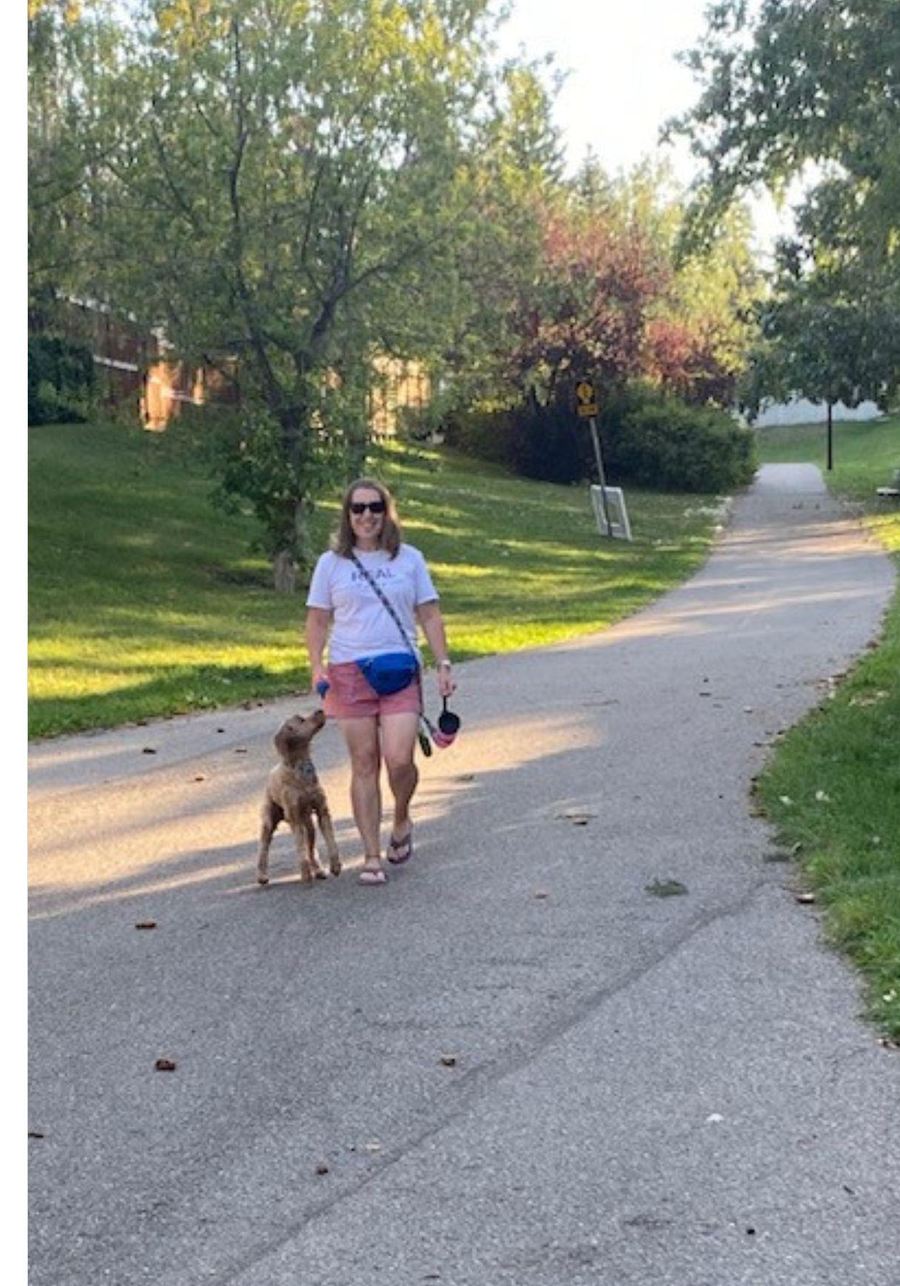Shauna is wearing a white t shirt, a blue fanny pack, and red shorts with purple flip flops. She is walking her dog on a leash in a residential neighbourhood.